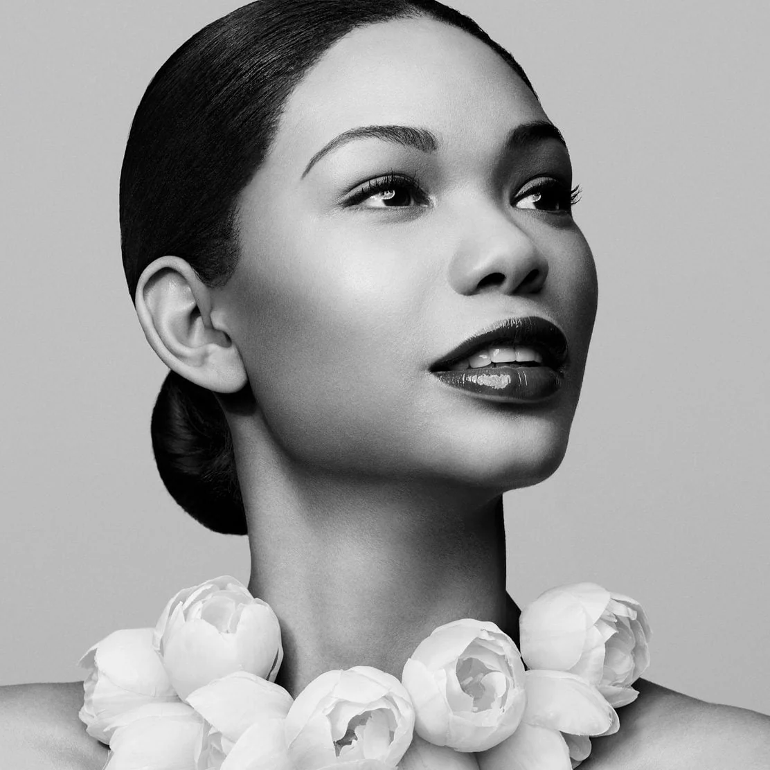 Black-and-white portrait of a woman with a sleek low bun and a collar of white peonies, gazing upward, for V.