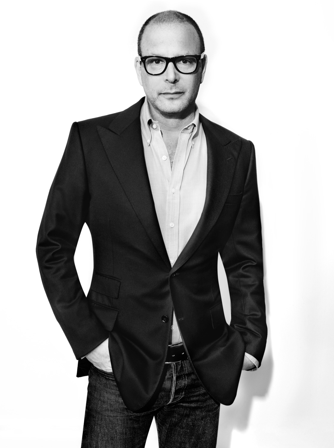 Black-and-white portrait of a man in thick glasses, dark blazer, open shirt and jeans, hands in pockets, for Elle.