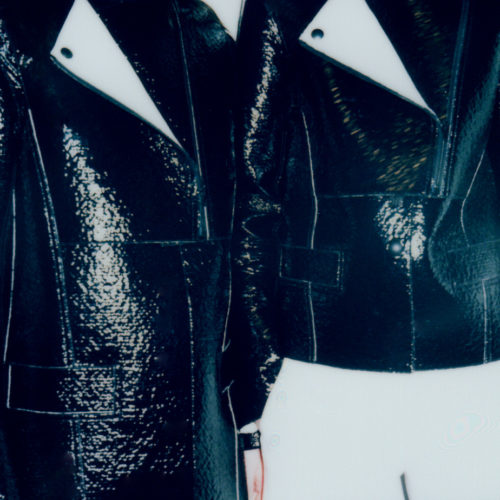 Two models in glossy black patent jackets with white piping and pointed lapels, cropped at the neck against a pale backdrop.