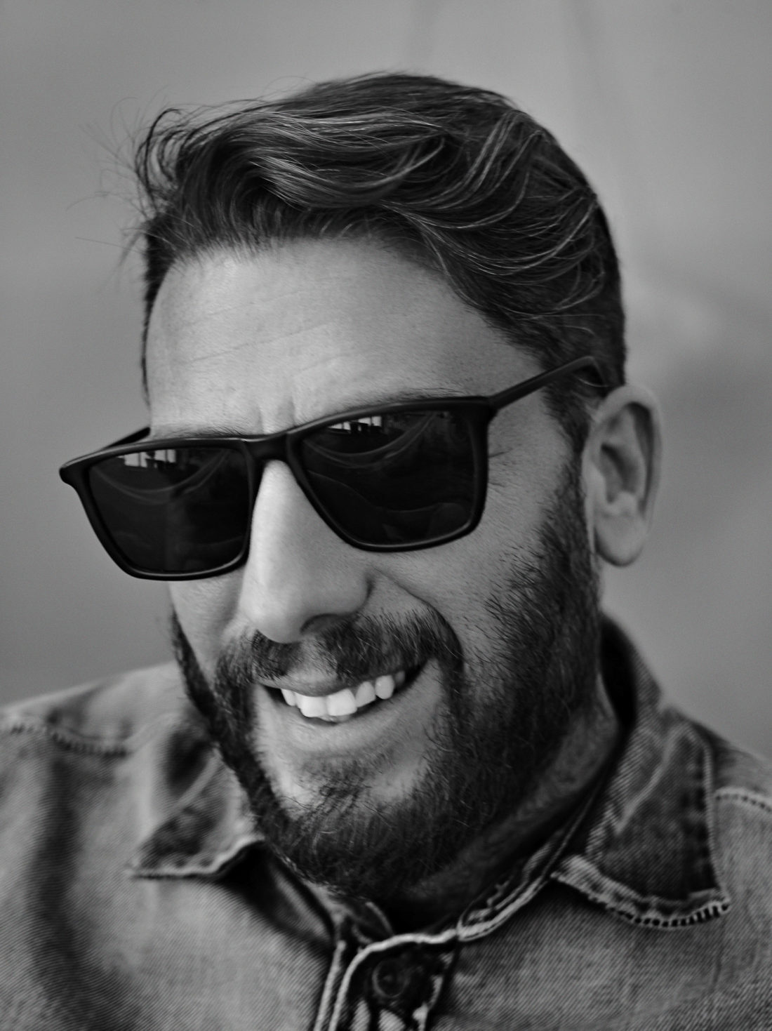 Black-and-white portrait of Andre Galvani smiling in dark sunglasses and a denim shirt, with a full beard.