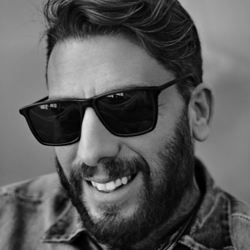 Black-and-white portrait of Andre Galvani smiling in dark sunglasses and a denim shirt, with a full beard.