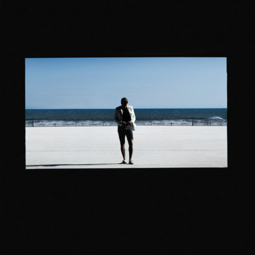 Brad Kroenig in a white jacket and shorts standing on a sunlit boardwalk before the ocean, framed by a black border.
