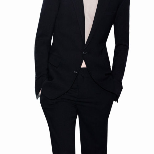 Model in a slim black two-button suit worn open over a bare chest, hands in pockets, on white, for Helmut Lang.