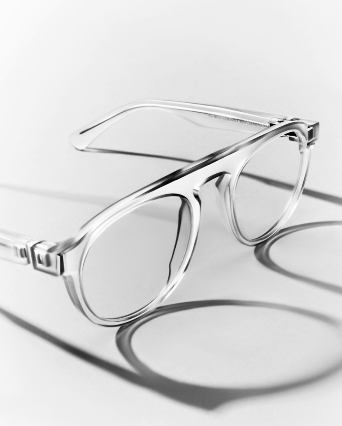 Clear acetate eyeglasses with metal hinges casting long shadows on a white surface, in black and white, for Maison Margiela.