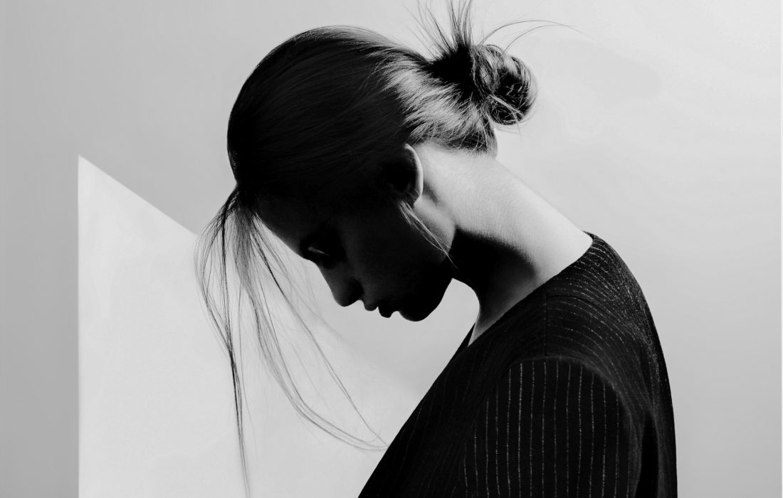 Black and white profile of a woman with head bowed, hair in a loose bun, wearing a dark pinstripe top against a pale wall.