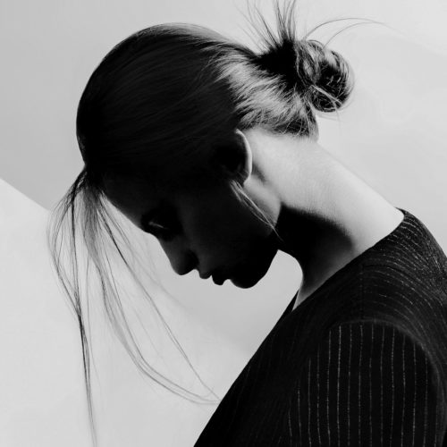 Black and white profile of a woman with head bowed, hair in a loose bun, wearing a dark pinstripe top against a pale wall.