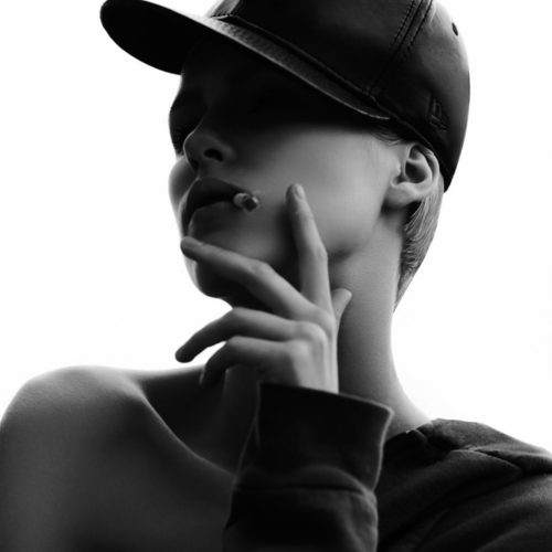 Black and white portrait of a woman in a leather baseball cap and off-shoulder sweatshirt, hand raised to a cigarette.