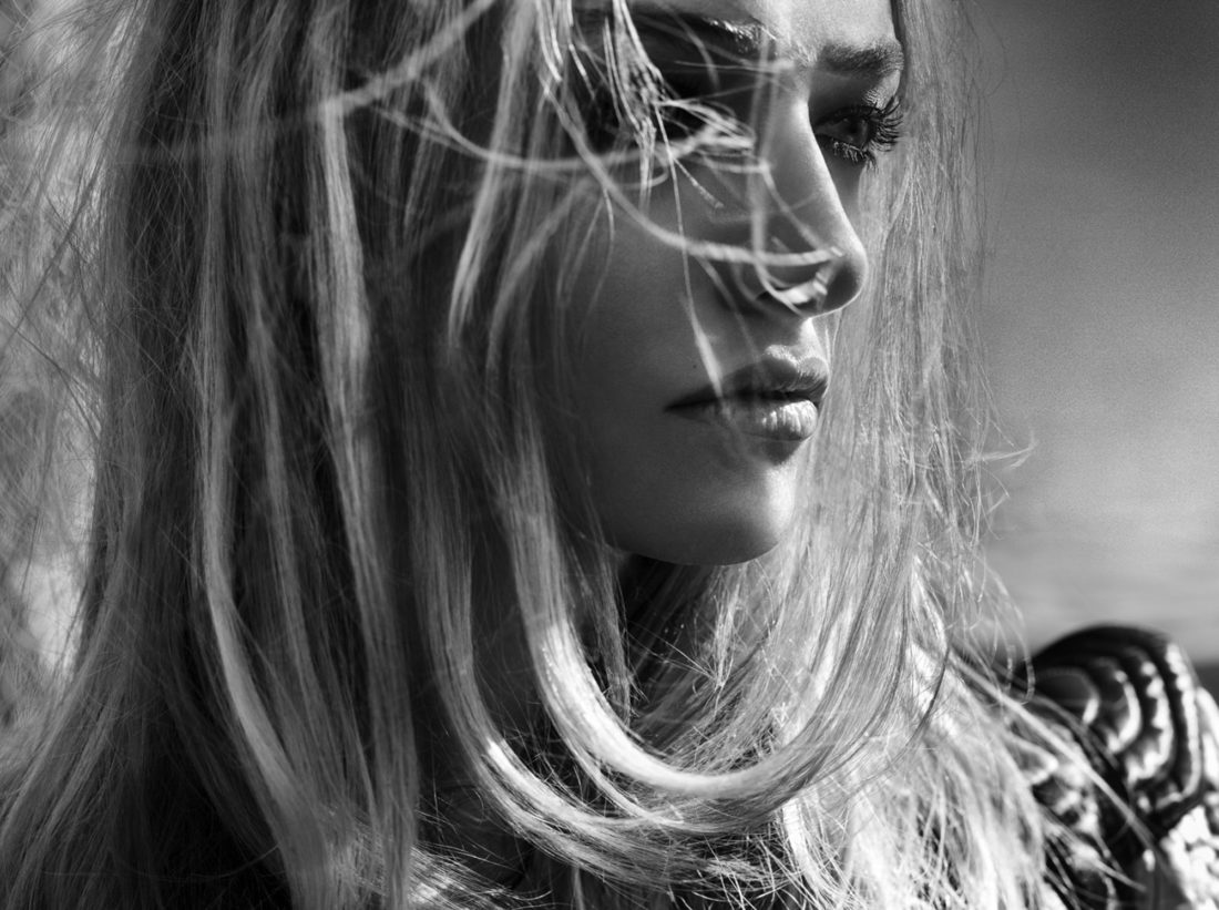 Black-and-white close-up of a woman in profile, long blonde hair blowing across her face, wearing a quilted jacket.