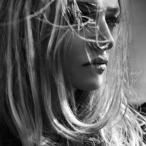 Black-and-white close-up of a woman in profile, long blonde hair blowing across her face, wearing a quilted jacket.