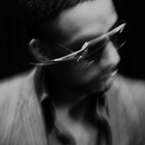 Black-and-white blurred portrait of Ryan Leslie in wraparound sunglasses and a pinstripe jacket against a dark background.