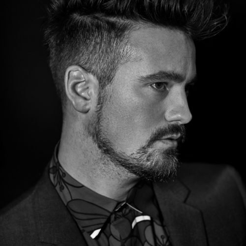 Black and white profile portrait of Sam Amoia with swept-back hair and beard, in a dark suit and floral patterned shirt.