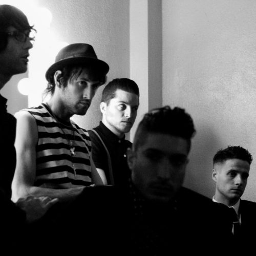 Black and white portrait of the five members of The Bravery gathered backstage beside a lit dressing-room mirror.