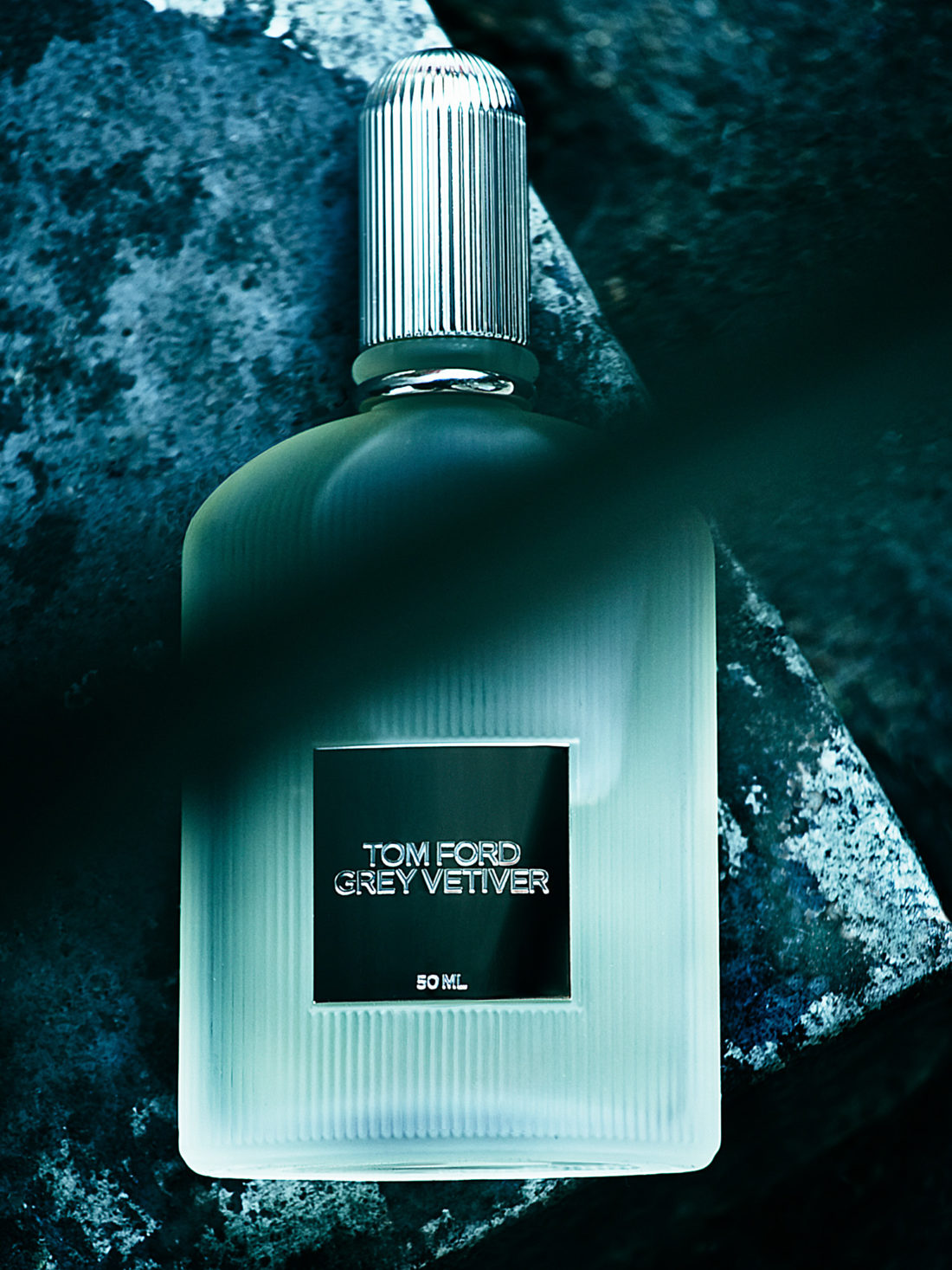 Frosted Grey Vetiver fragrance bottle with ribbed silver cap lying on dark blue-green stone, for Tom Ford.