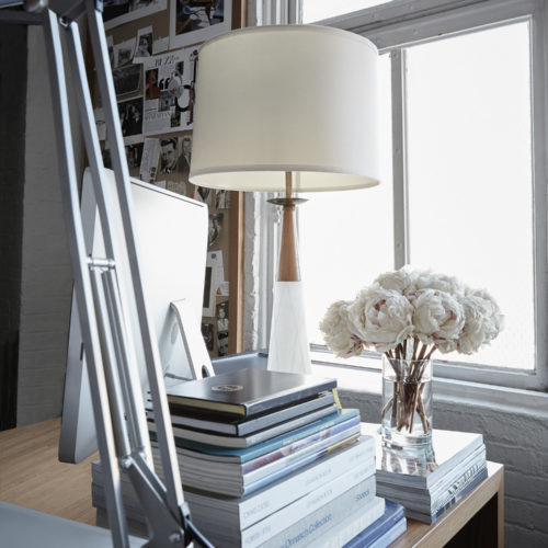 Desk by a loft window with a white table lamp, stacked books, a silver task lamp and a vase of white peonies, for Viana Art.