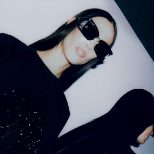 Model with long dark hair in oversized black sunglasses and black top, tilted flash shot backstage at fashion week, for The New York Times.