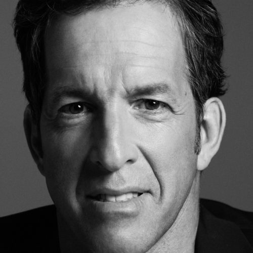 Black and white close-up portrait of designer Kenneth Cole in a dark jacket against a grey backdrop.