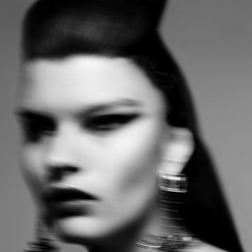 Black and white motion-blurred close-up of a model with sculpted dark hair, dark lips and large metallic earrings, for Sleek.