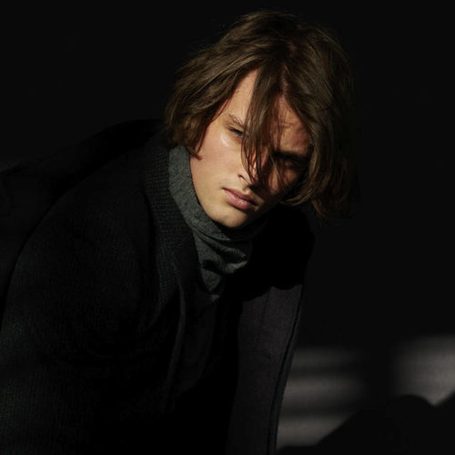 Young man with tousled brown hair in a dark coat and grey turtleneck, lit by a shaft of light against black, for VMAN.