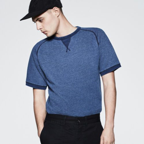 Male model in a heathered blue short-sleeve raglan sweatshirt, black cap and black trousers, for Saturdays NYC.
