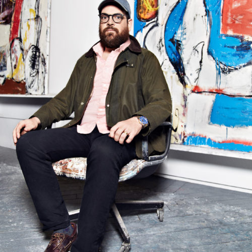 Painter Eddie Martinez seated in an office chair before his colourful abstract canvases in his studio, for Hercules.