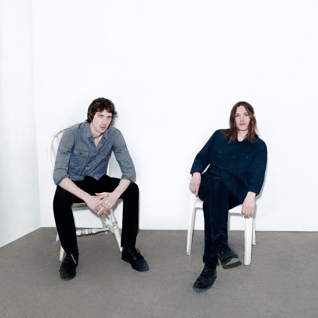 Two long-haired young men in dark shirts and black trousers seated on white chairs in a bare white studio, for The New York Times.