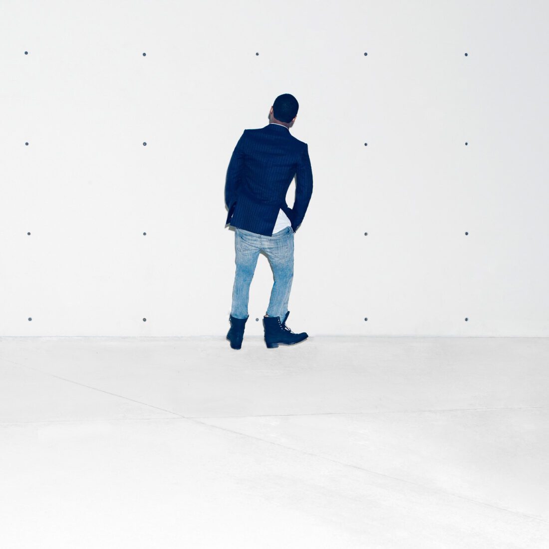 Man in navy pinstripe blazer, light jeans and boots stands facing a white concrete wall, for The New York Times.