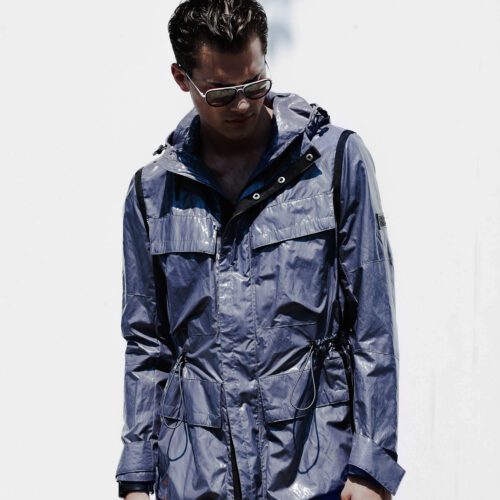 Man in aviator sunglasses wearing a shiny grey hooded nylon field jacket with drawstring waist against a white wall, for Out.
