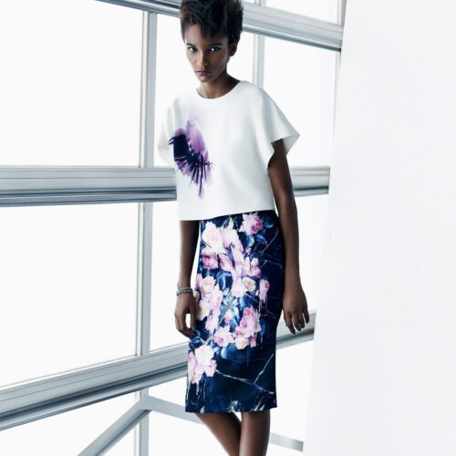 Model in a white boxy top with a painted eye and a navy rose-print pencil skirt by a bright window, for Saks Fifth Avenue.
