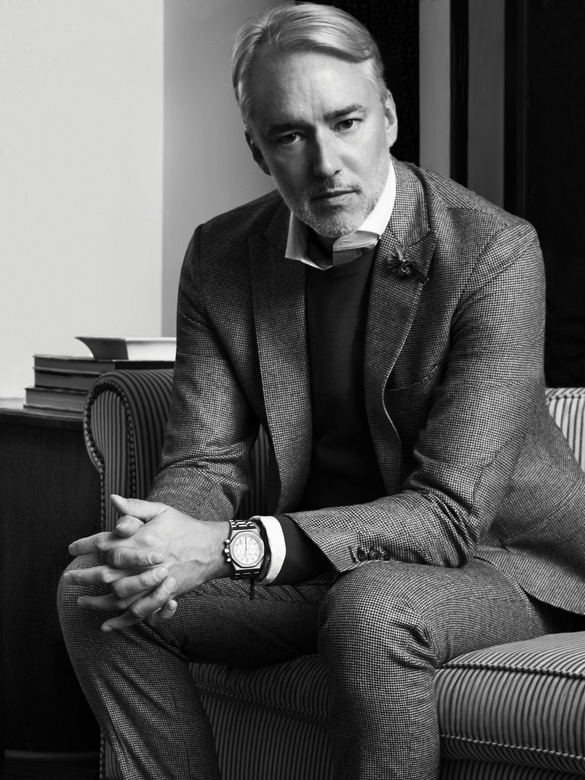 Black and white portrait of designer Michael Bastian in a tweed suit and wristwatch, seated on a striped armchair, for Hercules.