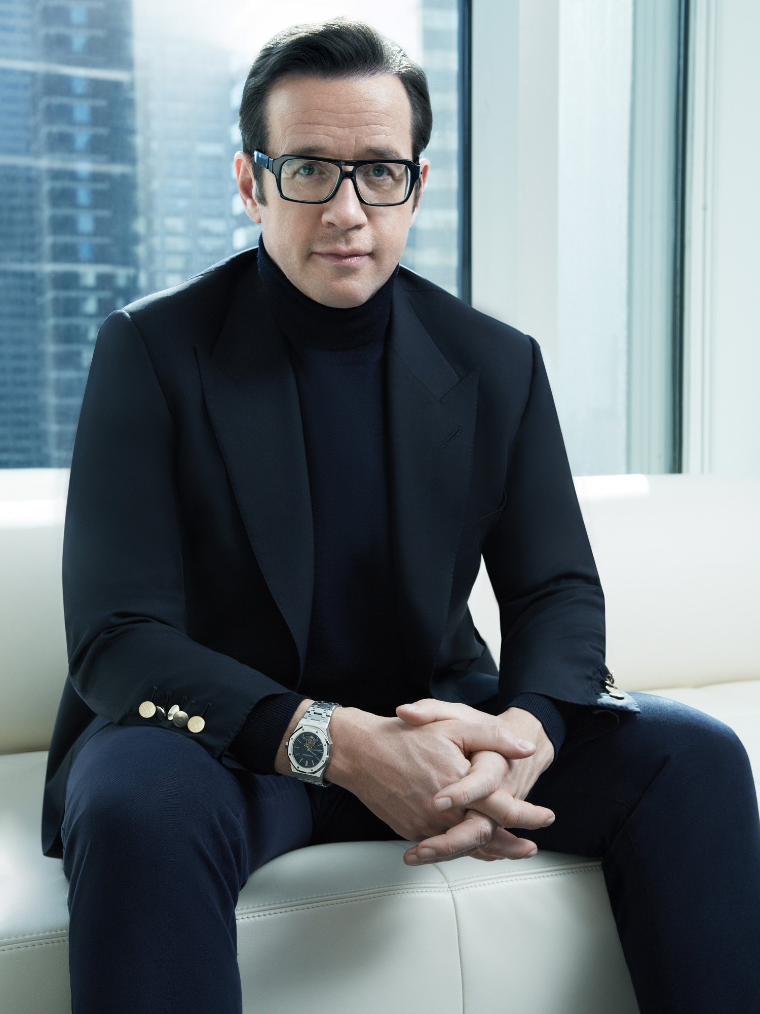François-Henry Bennahmias in glasses, black turtleneck and suit, seated on a white sofa by city windows, for Audemars Piguet.