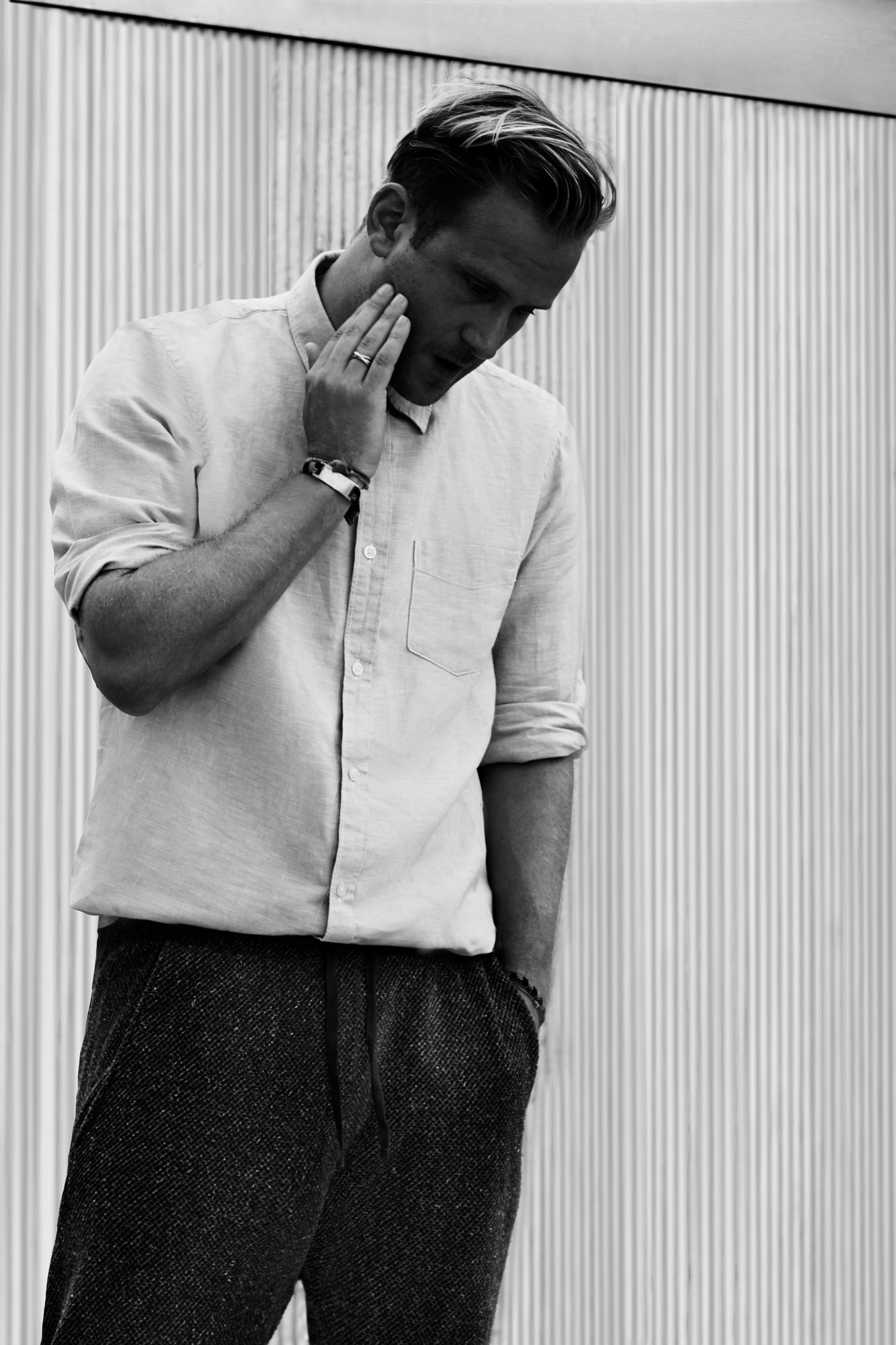 Black-and-white shot of a man in a rolled-sleeve linen shirt and tweed trousers by a corrugated wall, for Hercules.