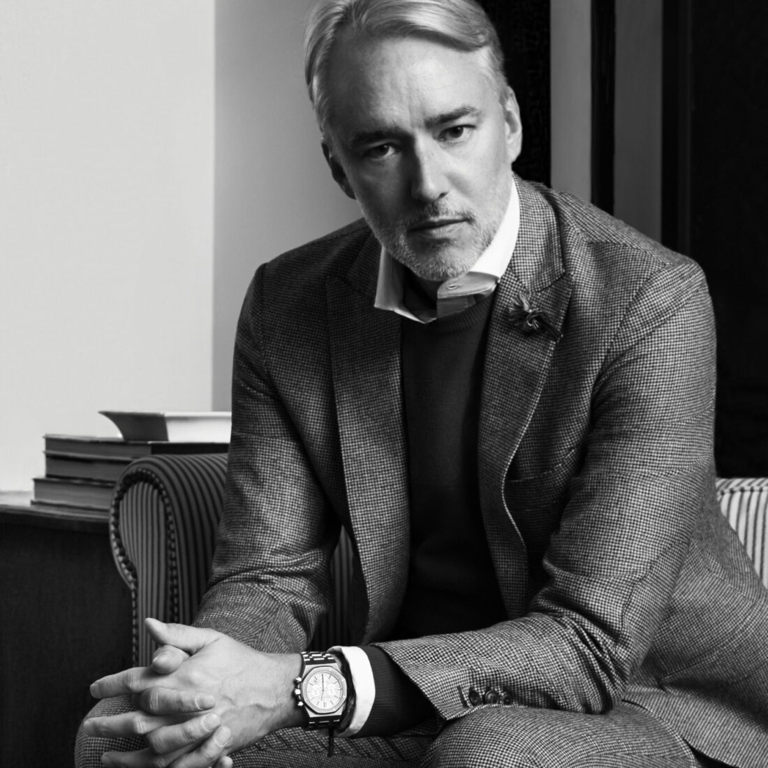 Black and white portrait of designer Michael Bastian in a houndstooth suit and chronograph watch, seated on a striped chair.