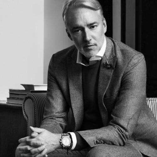 Black and white portrait of designer Michael Bastian in a houndstooth suit and chronograph watch, seated on a striped chair.