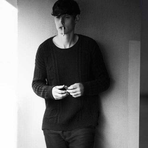 Black and white portrait of Adam Butcher in a dark cable-knit sweater, cigarette in mouth, holding a lighter against a wall.