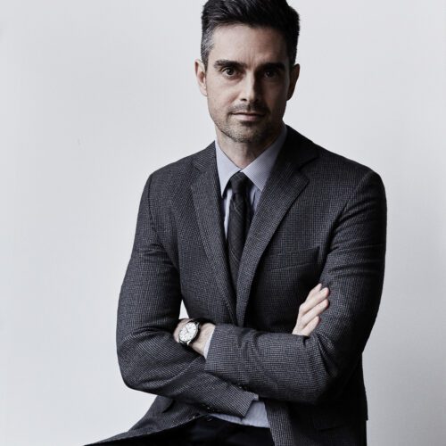 Andre Mellone in a grey checked blazer and dark tie, arms crossed, wearing a wristwatch against a pale grey wall.