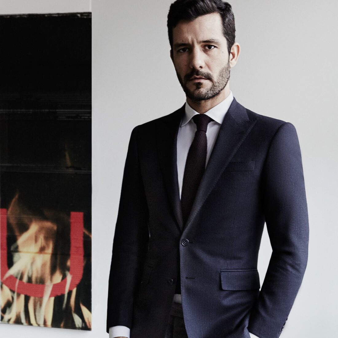 Andre Viana in a navy suit and dark knit tie, hand in pocket, beside a painting of flames on a white wall.
