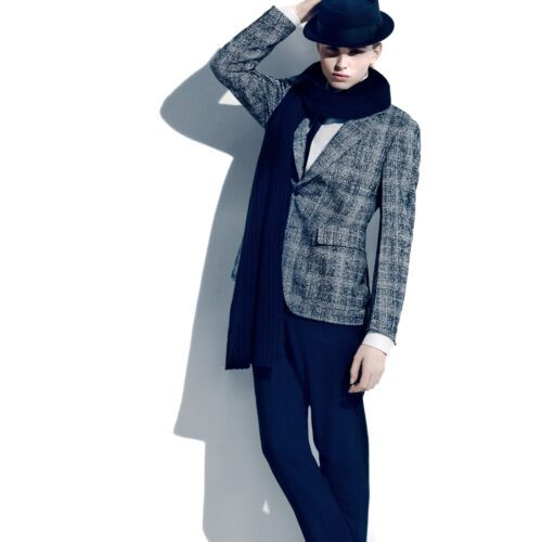 Model in a grey tweed blazer, navy fedora, long navy scarf and trousers posing against a white wall, for Apropos.