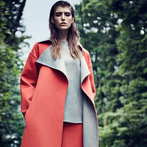 Model with slicked wet hair in a coral double-faced wool coat over a grey top, standing among green trees.