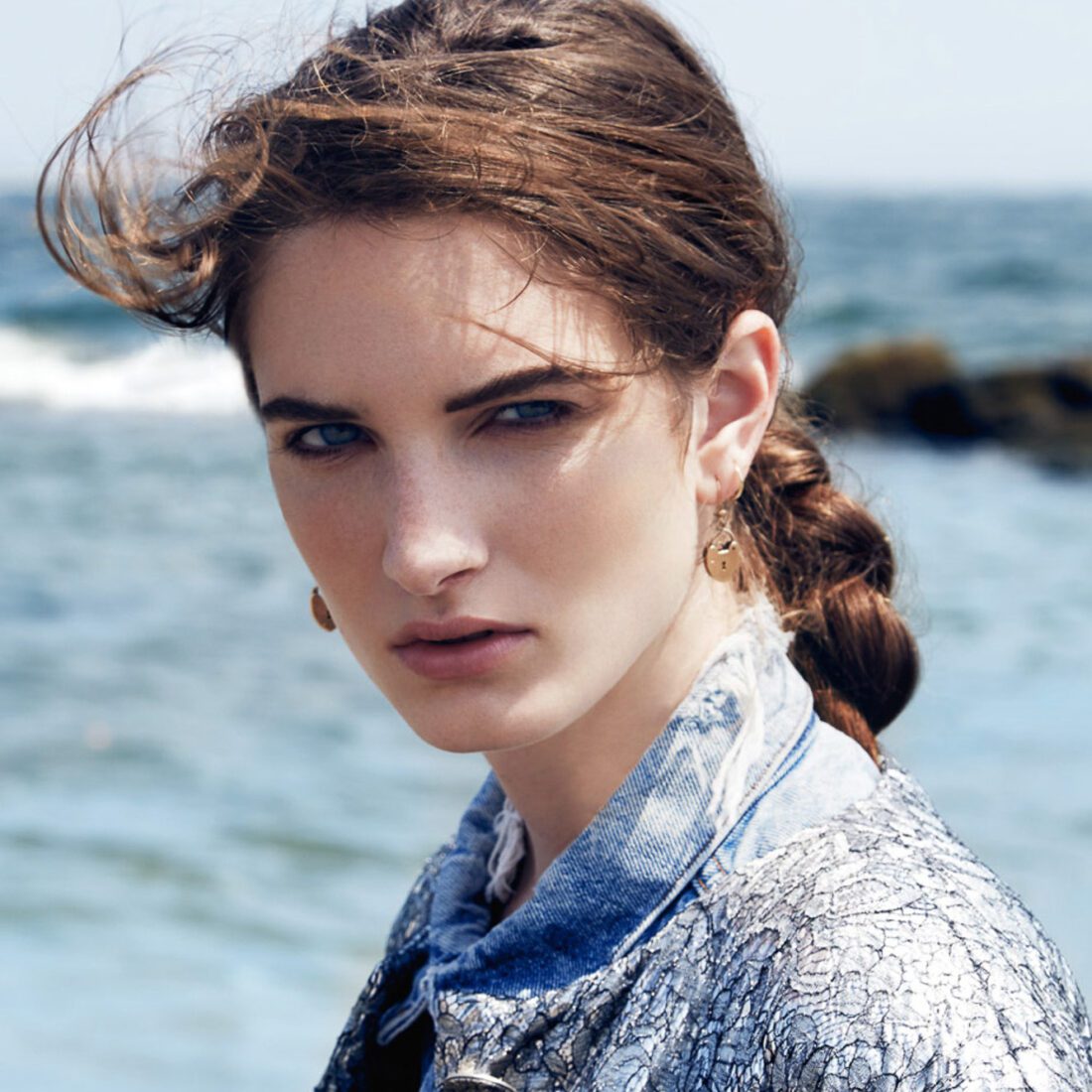 Carly Moore with a windswept braid, gold earrings and a denim and silver lace jacket against the ocean.
