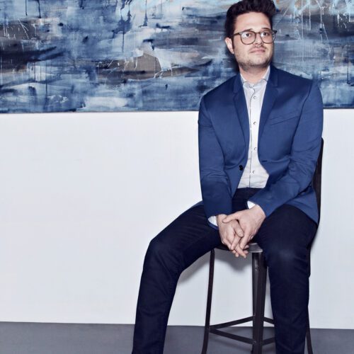 Cole Sternberg in glasses and a blue blazer seated on a metal stool beneath a large blue abstract painting.