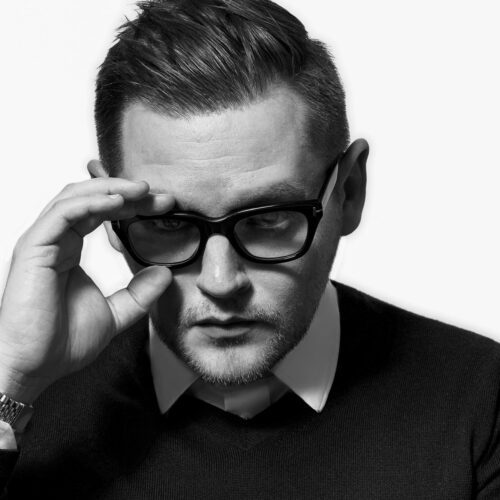 Black and white portrait of Conner Lutz adjusting thick black glasses, wearing a dark sweater over a white collared shirt.