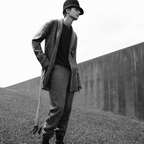 Black-and-white shot of a male model in a tweed cardigan, trousers and felt hat on grass beside a concrete wall.