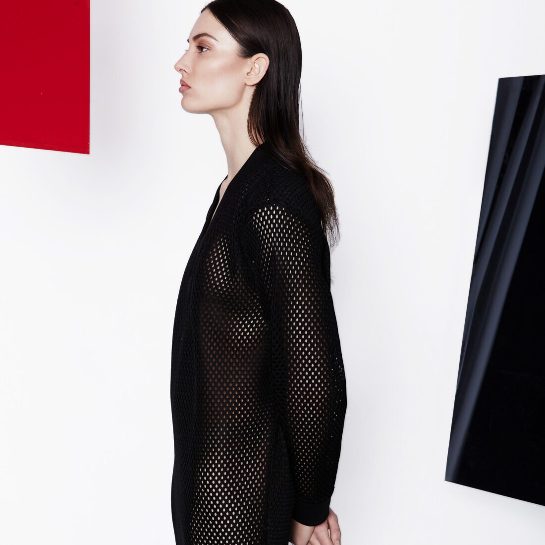 Model in profile wearing a sheer black mesh cardigan against a white wall with red and black panels.