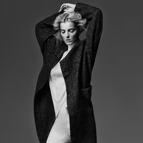 Black-and-white portrait of model Denisa Dvorakova in a long dark tweed coat and white shirt, hands raised to her hair.