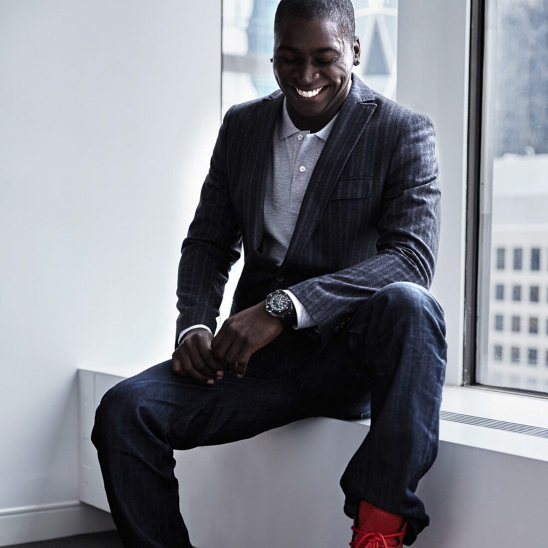 Didi Gregorius smiling on a window ledge in a pinstripe blazer, grey polo, dark jeans, watch and red sneakers.