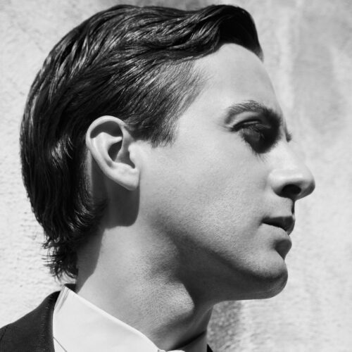 Black and white profile portrait of Eddie Borgo with slicked-back hair, white collar and dark jacket against a textured wall.