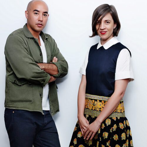 Suno designers Max Osterweis and Erin Beatty against a white wall, in olive jacket and printed skirt, for Elle.