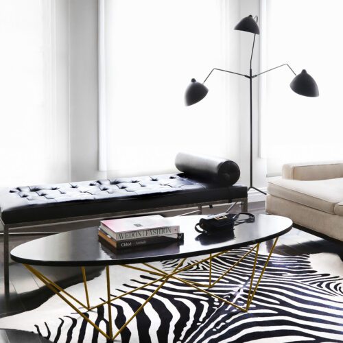 Living room with black tufted daybed, oval coffee table on gold legs, zebra-hide rug and three-arm lamp, for Harper's Bazaar.