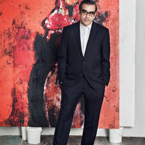 Artist Enoc Perez in a dark suit and glasses standing before a large red abstract painting in his studio, for Hercules.