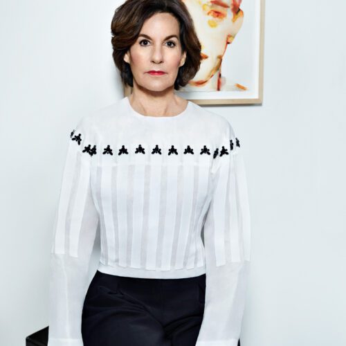 Kim McCarty seated in a white pleated blouse with black beading, a framed watercolour behind her, for Hercules.
